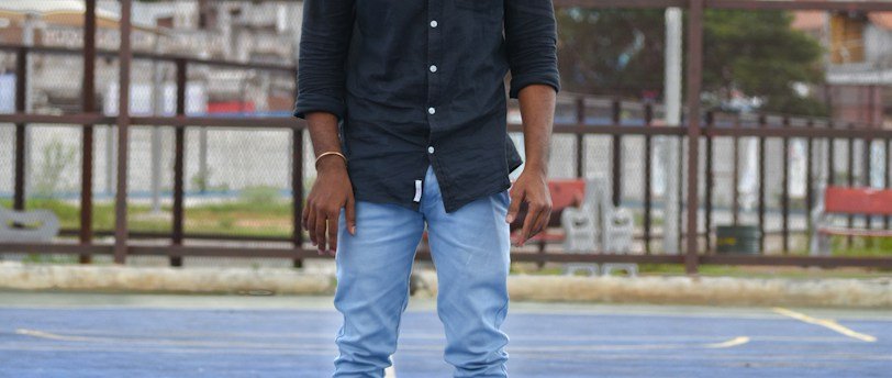 A person stands on an outdoor basketball court with a basketball positioned in front of their head, obscuring their face. They are wearing a black shirt, light blue jeans, and sandals. In the background, a basketball hoop and urban buildings are visible.