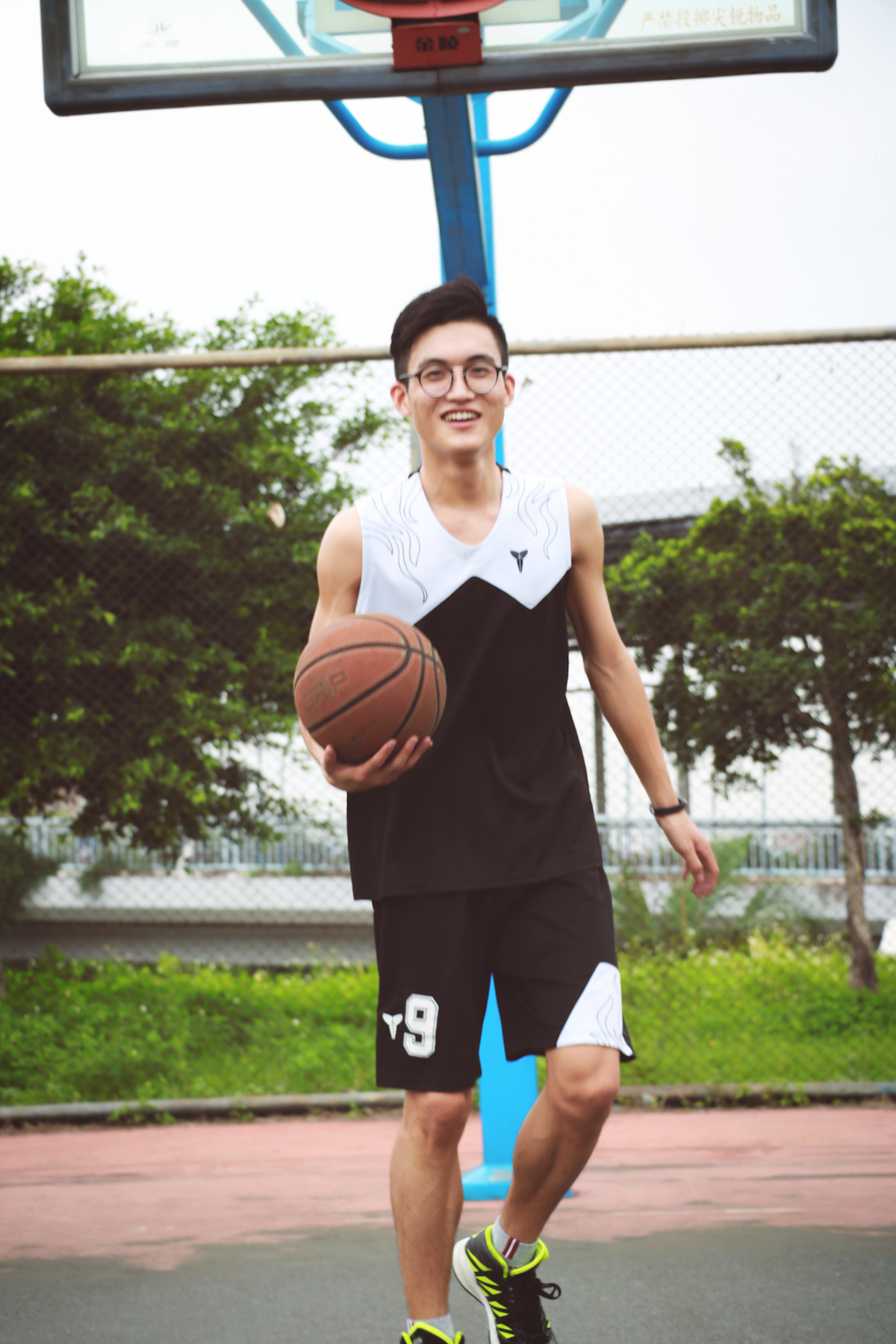 A person with glasses, wearing a black and white sports outfit is playing basketball on an outdoor court. He holds a basketball with one hand while smiling and walking towards the camera. The background includes a basketball hoop and greenery.