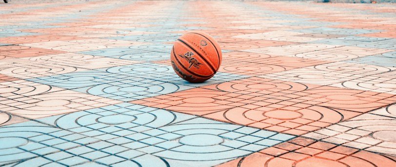 A basketball rests in the center of a colorful patterned courtyard. The surface is composed of red and blue tiles with intricate designs. In the background, a few people are visible near playground equipment and buildings.